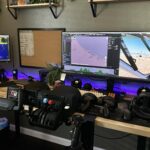 Home flight simulator cockpit setup with throttle quadrant centre-foreground, yoke, dual ultrawide monitor displays running Microsoft Flight Simulator, mixed HOTAS controls, purple LED backlighting
