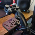 MOZA AB9 force feedback flight stick base with MH16 fighter-style grip clamped to a wooden desk, Corsair speaker and bookshelf visible in soft focus