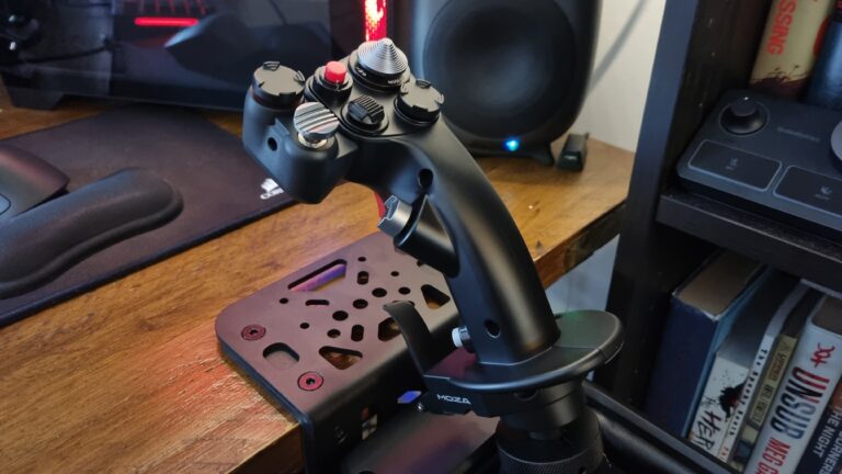 MOZA AB9 force feedback flight stick base with MH16 fighter-style grip clamped to a wooden desk, Corsair speaker and bookshelf visible in soft focus