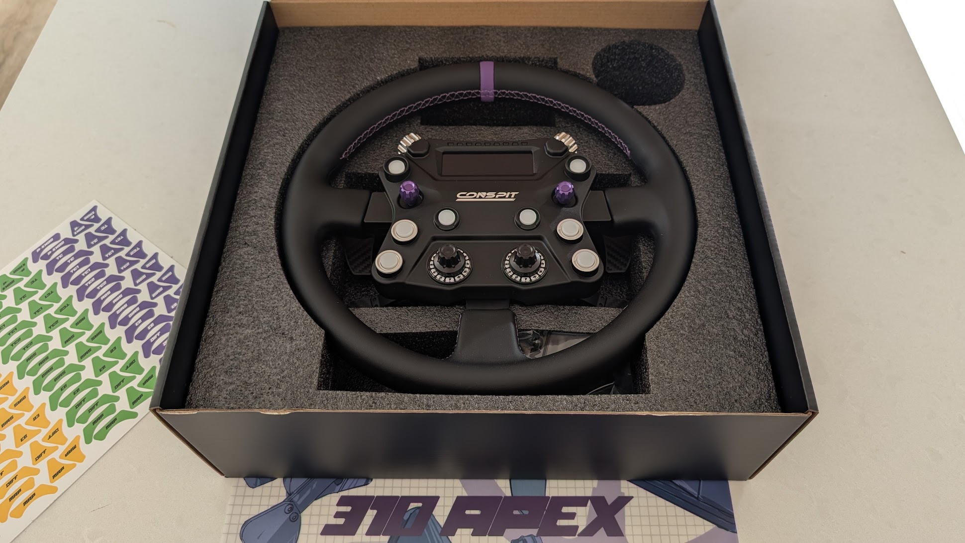 Conspit 310 APEX box open showing the wheel in foam, the sticker sheet and the illustrated art sheet