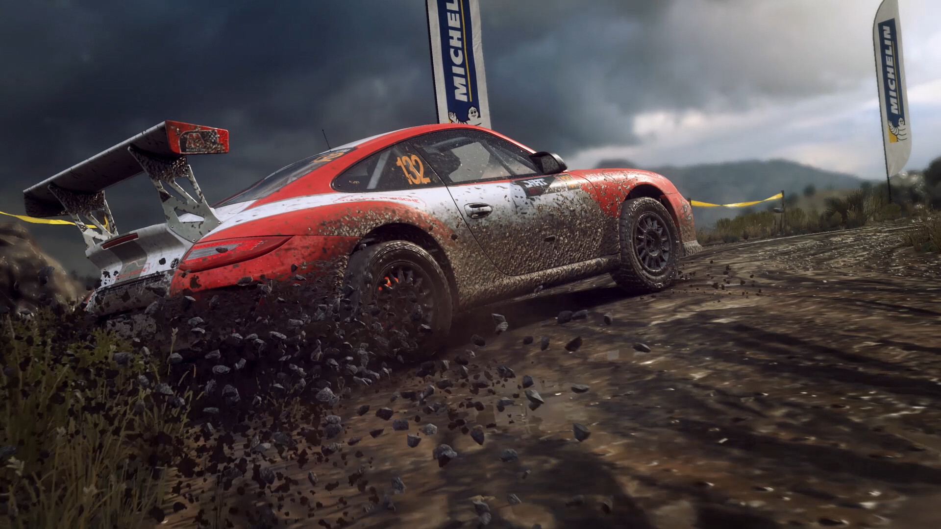 Red and white Porsche 911 rally car throwing up gravel past Michelin banners in DiRT Rally 2.0