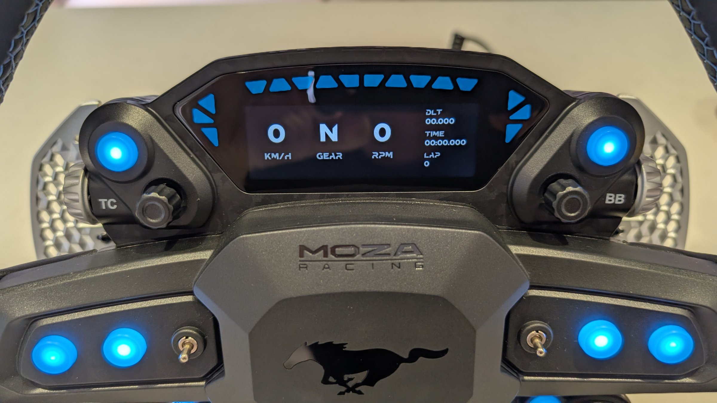 Close-up of the MOZA Mustang GTD wheel display reading 0 km/h, gear N and 0 RPM, with blue rev lights and the TC and BB labels