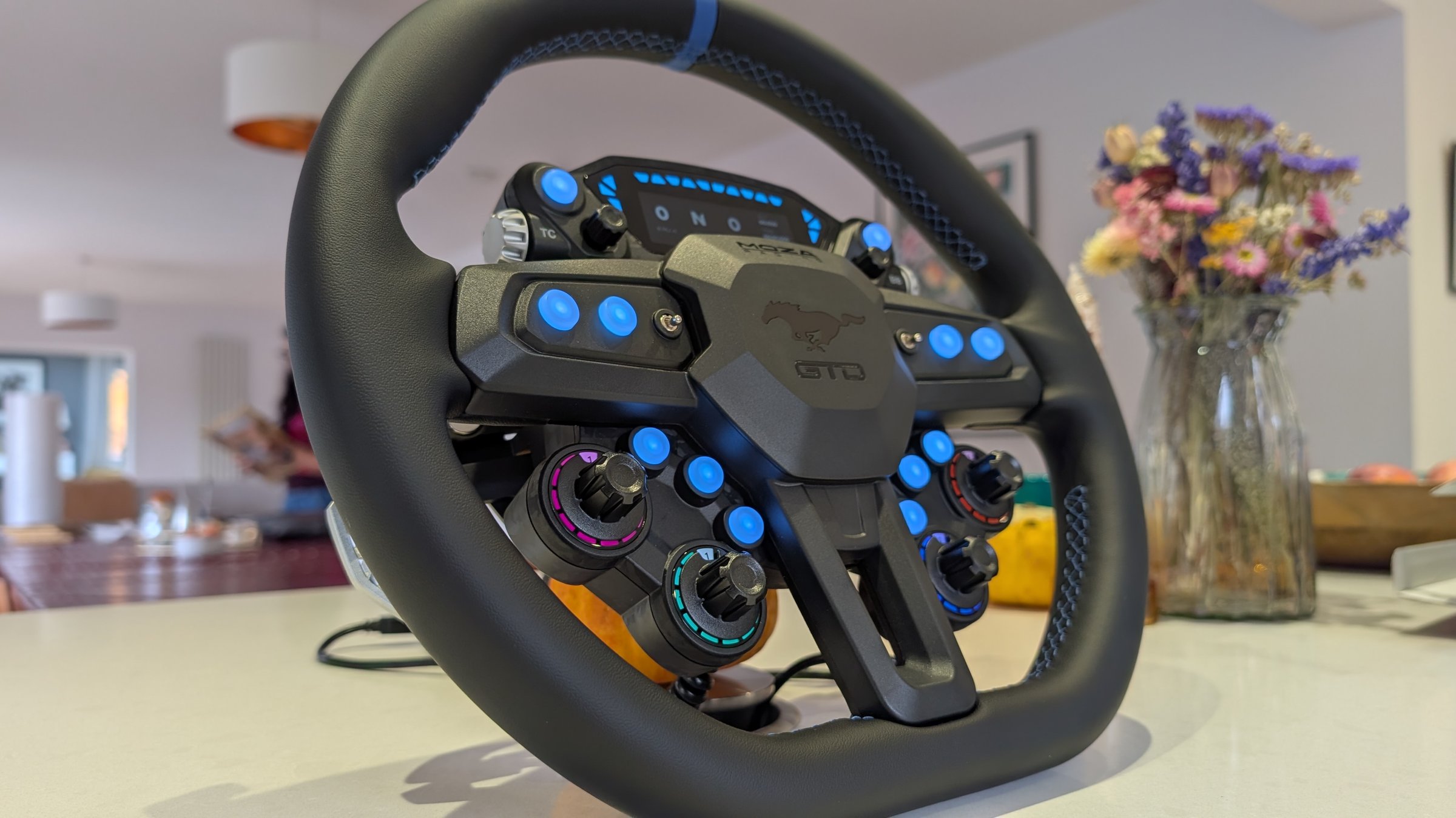 MOZA Mustang GTD wheel from a low side angle, showing the rotary encoders with coloured LED rings and the stitched rim