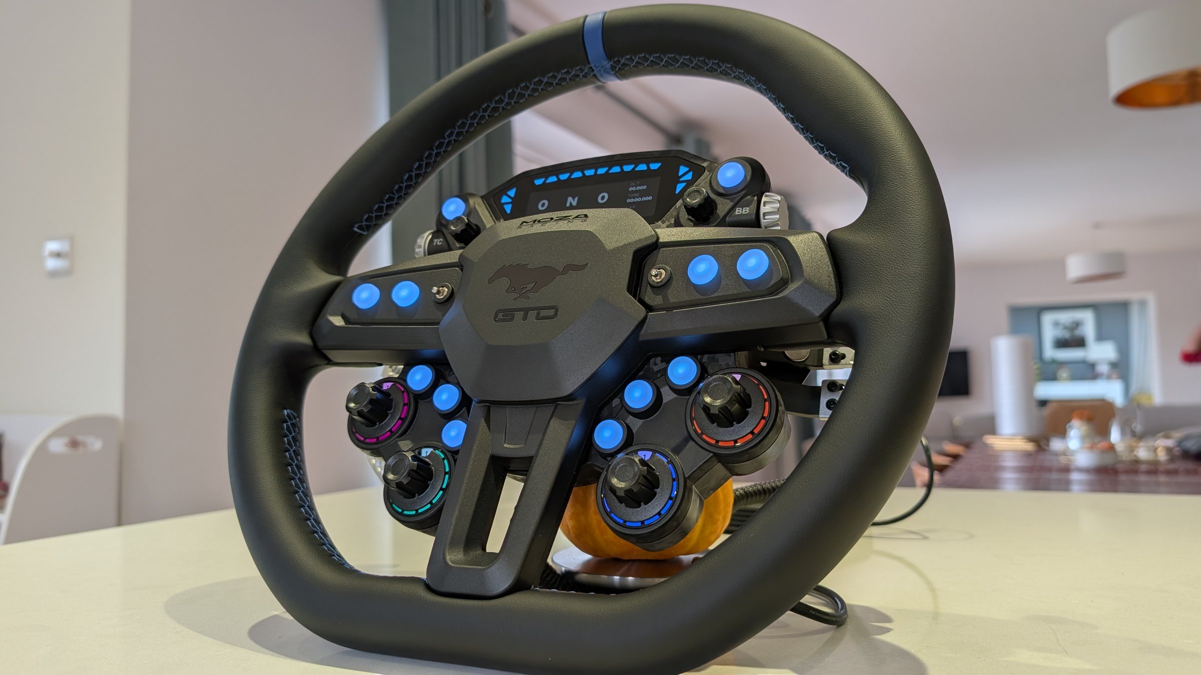 Low three-quarter view of the MOZA Mustang GTD wheel showing the blue top stripe, blue stitching and the lit controls